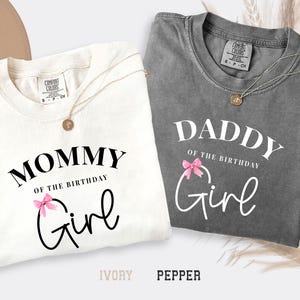 May include: Two t-shirts, one ivory and one pepper, with the text "Mommy of the Birthday Girl" and "Daddy of the Birthday Girl" in white script. Each shirt has a pink bow graphic. Gold necklaces are draped on the shirts.
