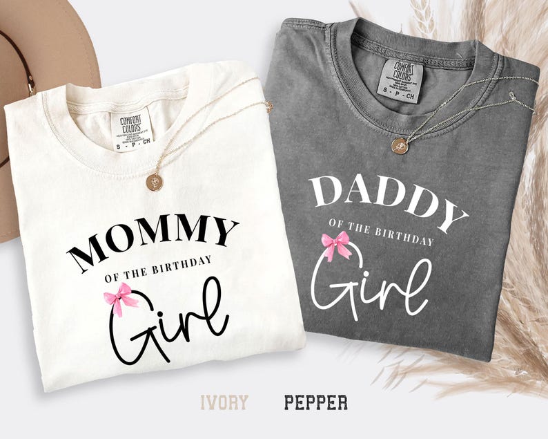 May include: Two t-shirts, one ivory and one pepper, with the text "Mommy of the Birthday Girl" and "Daddy of the Birthday Girl" in white script. Each shirt has a pink bow graphic. Gold necklaces are draped on the shirts.