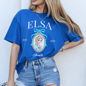May include: A royal blue t-shirt featuring the name "ELSA" in white script above a portrait of Elsa from the movie Frozen. The shirt includes the text "EST. 2013" and "Arendelle" in a decorative font. The shirt is made of a soft material.