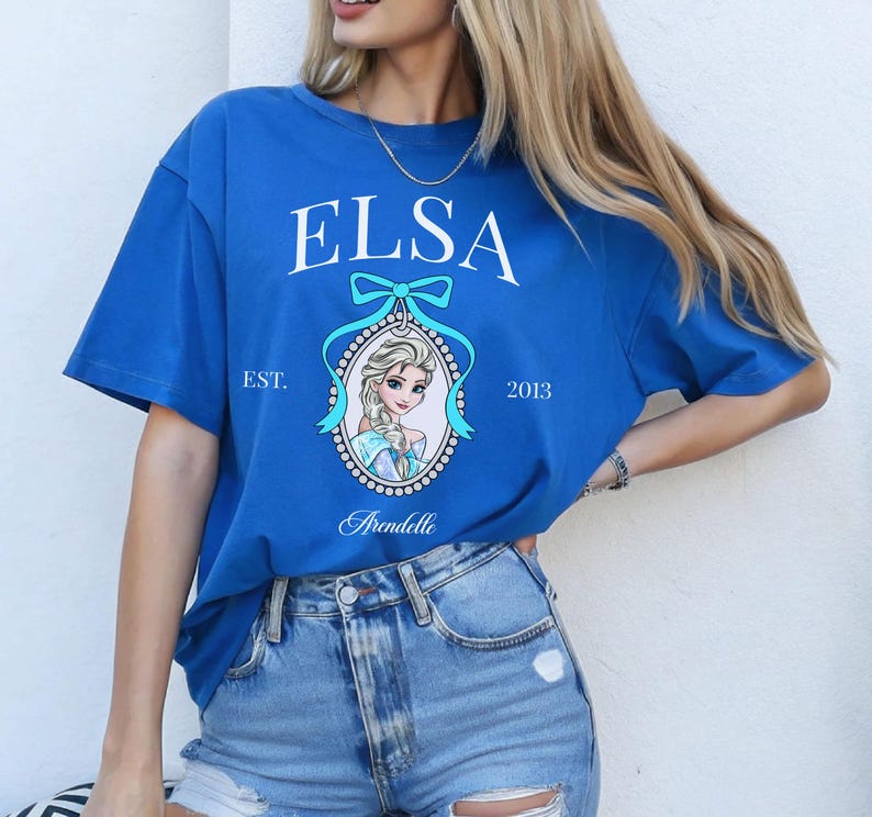 May include: A royal blue t-shirt featuring the name "ELSA" in white script above a portrait of Elsa from the movie Frozen. The shirt includes the text "EST. 2013" and "Arendelle" in a decorative font. The shirt is made of a soft material.