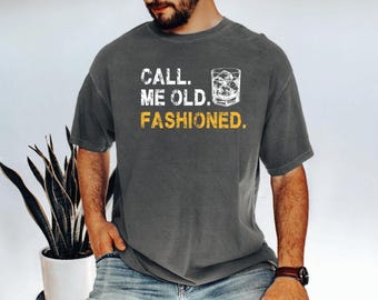 May include: Dark gray t-shirt with the text "CALL. ME OLD. FASHIONED." in white and yellow. A graphic of a cocktail glass with ice is printed on the shirt. The shirt is a casual wear item.