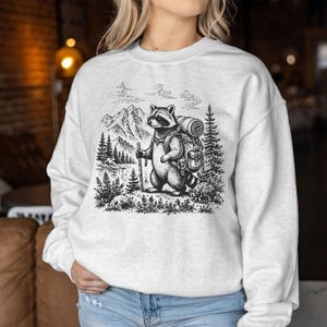May include: A light heather gray crewneck sweatshirt featuring a black ink-style graphic of a raccoon hiking in the mountains. The raccoon wears a backpack, carries a walking stick, and is surrounded by pine trees, a lake, and rocky peaks.