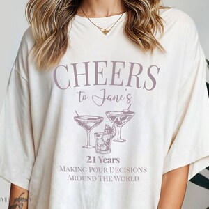 Peut inclure: T-shirt blanc avec le texte "Cheers to Jane's 21 Years Making Pour Decisions Around The World" et un graphique de trois verres &agrave; martini.