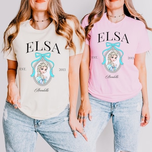 May include: Two short-sleeved t-shirts, one cream and one pink, each featuring the name "ELSA" in black, above a portrait of a cartoon character. The character is framed by a light blue bow and a decorative border. The word "Arendelle" is below the portrait.