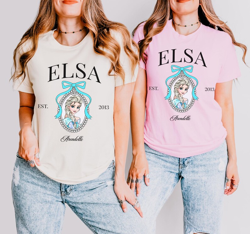 May include: Two short-sleeved t-shirts, one cream and one pink, each featuring the name "ELSA" in black, above a portrait of a cartoon character. The character is framed by a light blue bow and a decorative border. The word "Arendelle" is below the portrait.