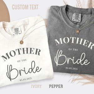 May include: Ivory and pepper-colored t-shirts with the text "MOTHER OF THE Bride" and the date "05.05.2025" printed on the front. Each shirt has a gold-colored necklace. The shirts are labeled "IVORY" and "PEPPER".