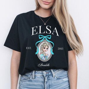 May include: Black t-shirt featuring the name "ELSA" in white, above a portrait of a cartoon character. The portrait is framed with a light blue bow and a dotted border. The text "EST. 2013" and "Arendelle" are also present.
