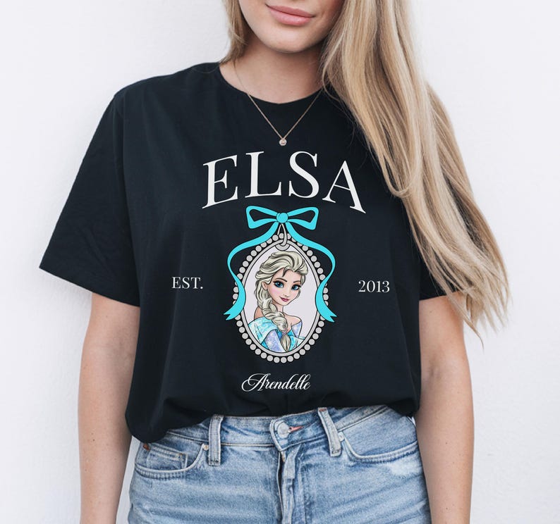 May include: Black t-shirt featuring the name "ELSA" in white, above a portrait of a cartoon character. The portrait is framed with a light blue bow and a dotted border. The text "EST. 2013" and "Arendelle" are also present.