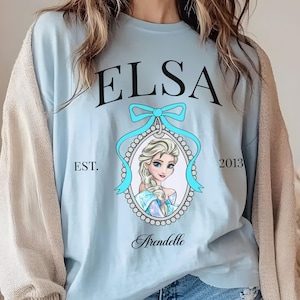 May include: Light blue sweatshirt featuring the name "ELSA" in black script above a portrait of a blonde woman with a blue bow and the word "Arendelle" below. The text "EST. 2013" is also visible.