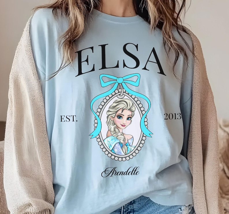 May include: Light blue sweatshirt featuring the name "ELSA" in black script above a portrait of a blonde woman with a blue bow and the word "Arendelle" below. The text "EST. 2013" is also visible.