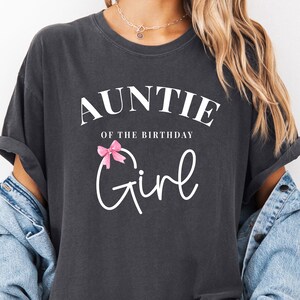 May include: Dark gray t-shirt with the text "AUNTIE of the BIRTHDAY Girl" in white script. A pink bow graphic is above the word "Girl". The shirt has rolled-up sleeves and is worn with a denim jacket.