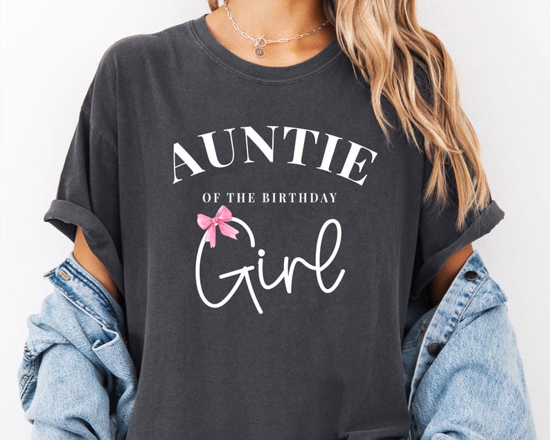 May include: Dark gray t-shirt with the text "AUNTIE of the BIRTHDAY Girl" in white script. A pink bow graphic is above the word "Girl". The shirt has rolled-up sleeves and is worn with a denim jacket.