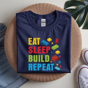 May include: Navy blue t-shirt with the words "EAT SLEEP BUILD REPEAT" in colorful block letters and block graphics. The shirt is displayed on a woven surface with a pair of white sneakers visible in the background.