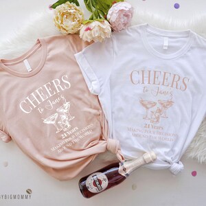 Peut inclure: Deux t-shirts de couleur claire avec le texte "CHEERS to Jane's 21 Years MAKING POUR DECISIONS AROUND THE WORLD" et un graphique de deux verres &agrave; martini.