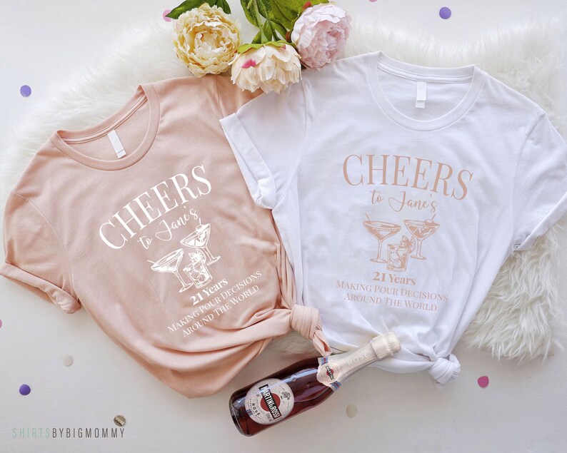 Peut inclure: Deux t-shirts de couleur claire avec le texte "CHEERS to Jane's 21 Years MAKING POUR DECISIONS AROUND THE WORLD" et un graphique de deux verres &agrave; martini.