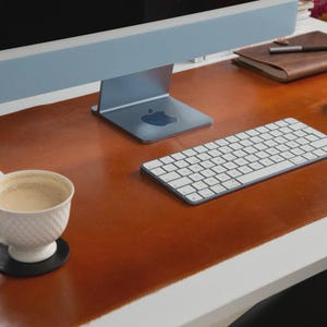 May include: A desk with a brown leather mat, a computer monitor, keyboard, and mouse. A white smartphone and a leather notebook are also on the desk. A cup of coffee sits on a black coaster.