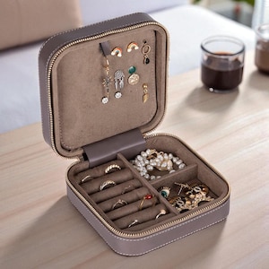 May include: Open gray jewelry box with a gold zipper, filled with various earrings, rings, and necklaces. The interior is lined with a soft, light brown material. The box is square and has a stitched border.