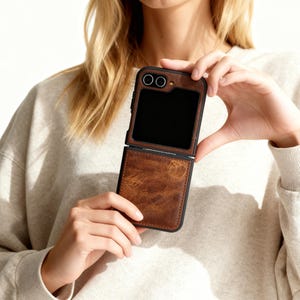 May include: A brown leather phone case with a black frame, open to reveal a black screen. The case has a textured appearance and a contrasting black border. The phone case is held in front of a neutral background.
