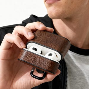 Pode incluir: Uma capa de couro castanho para AirPods com acabamento preto e um mosquetão preto. A capa está aberta, revelando AirPods brancos. O couro tem uma aparência texturizada.