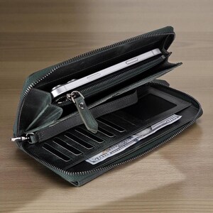 Puede incluir: Cartera de cuero verde oscuro abierta con cierre de cremallera. La cartera está abierta, revelando un teléfono inteligente, ranuras para tarjetas de crédito y un compartimento para billetes con un montón de billetes. La cartera está sobre una superficie de madera.