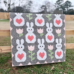 May include: A handmade quilt featuring a repeating pattern of white bunnies with red hearts, pink tulips, and green stems on a gray background. The quilt has a pink border and is displayed outdoors on a wooden fence.