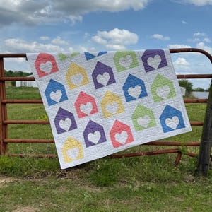 May include: A handmade quilt featuring a repeating pattern of colorful house shapes, each with a heart-shaped cutout. The houses are in shades of pink, yellow, purple, green, and blue, set against a white background. The quilt is draped over a rusty metal fence.