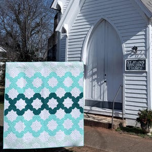 May include: A handmade quilt with a repeating geometric pattern in white, teal, and dark teal. The quilt is displayed outdoors in front of a white church. The quilt's design features interlocking shapes, creating a visually appealing textile.