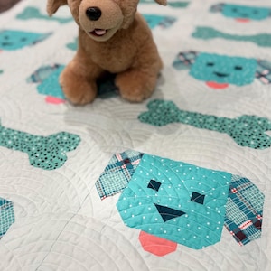 May include: A light blue and white quilt with a patchwork design of dogs and bones. The quilt is quilted with a wavy pattern. A brown plush dog toy is sitting on the quilt.