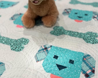 May include: A light blue and white quilt with a patchwork design of dogs and bones. The quilt is quilted with a wavy pattern. A brown plush dog toy is sitting on the quilt.