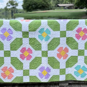 May include: A handmade quilt with a floral pattern. The quilt features large, colorful flowers in shades of pink, purple, peach, and teal, set against a green and white background. The quilt is displayed outdoors, with a blurred natural backdrop.