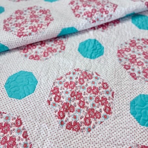 May include: Close-up of a quilted fabric with a white background and a pattern of pink floral circles and turquoise geometric shapes. The fabric has a dotted pattern and a quilted texture.