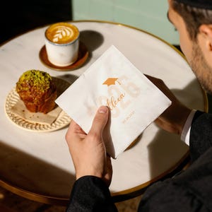 May include: A white square napkin with a graduation cap graphic and the name "Alexa" printed in a soft orange color. The date "03.20.2023" is also printed on the napkin. A cup of coffee and a pastry are on the table.