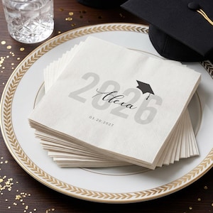 May include: A stack of white paper napkins with a graduation theme. The napkins are printed with the year "2026", the name "Alexa", and a graduation cap icon. The date "03.20.2027" is also printed on the napkins. The napkins are on a white plate with a gold rim.