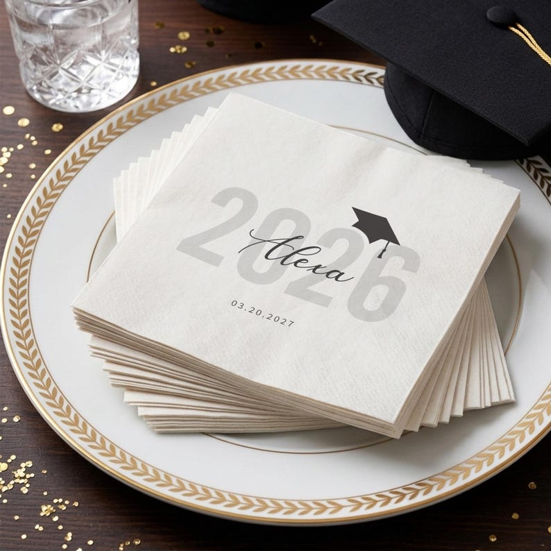May include: A stack of white paper napkins with a graduation theme. The napkins are printed with the year "2026", the name "Alexa", and a graduation cap icon. The date "03.20.2027" is also printed on the napkins. The napkins are on a white plate with a gold rim.
