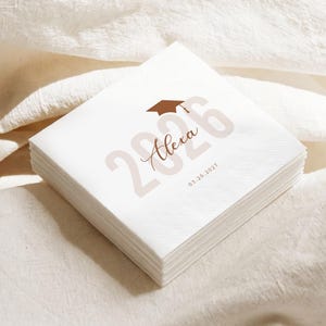 May include: A stack of white paper napkins with a graduation cap graphic and the text "2026 Alexa 03.20.2027" printed in brown. The napkins are stacked neatly on a white surface.