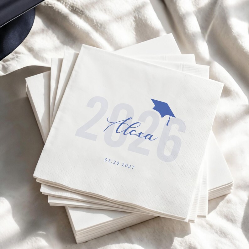 May include: A stack of white paper napkins with blue text and a graduation cap graphic. The text includes the year "2026", the name "Alexa", and the date "03.20.2027". The napkins are stacked on a white surface.