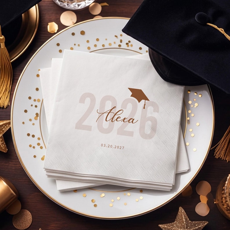 May include: White graduation napkins with the name "Alexa" and the year "2026" printed in a soft brown color. A graduation cap graphic is above the year. The napkins are stacked on a white plate with gold trim and gold confetti.