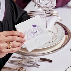 May include: A white graduation napkin with the name "Alexa", the year "2026", and a purple graduation cap graphic. The napkin is held near a champagne glass and silverware on a table setting. The date "03.20.2027" is printed on the napkin.