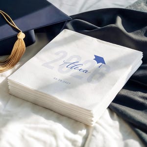 May include: A stack of white paper napkins with the text "2026 Alexa" and a graduation cap graphic in blue. The date "03.20.2027" is also printed on the napkins. A navy blue graduation cap with a gold tassel is in the background.