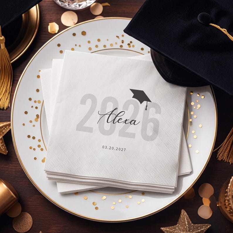 May include: A stack of white paper napkins with the text "2026 Alexa" and a graduation cap graphic. The date "03.20.2027" is printed below. The napkins are on a white plate with gold trim, with graduation caps and gold confetti in the background.