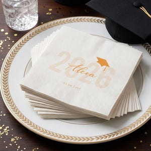 May include: A stack of off-white graduation napkins with gold text and a mortarboard graphic. The text reads "2026 Alexa 03.20.2027". The napkins are on a white plate with a gold leaf design.