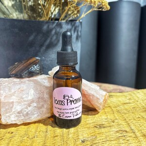 May include: A small amber glass bottle with a black dropper, labeled "Texas Prema" in pink. The bottle sits on a wooden surface, with pink and brown stones and a black pot with dried flowers in the background.