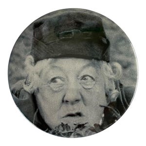 May include: A circular pinback button featuring a black and white photographic portrait of an older individual with curly hair wearing a dark cap. The person has a wide-eyed, surprised expression with their gaze directed toward the side.