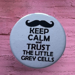 May include: A circular white pinback button featuring a black graphic of a mustache at the top. Below the mustache, the text reads: KEEP CALM and TRUST THE LITTLE GREY CELLS in bold black capital letters, centered on the surface of the round button.
