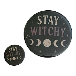 May include: Two circular black pin-back buttons of different sizes featuring the white text STAY WITCHY in a bold font. Below the text, a series of five moon phases are depicted in white, ranging from a crescent to a full moon and back.