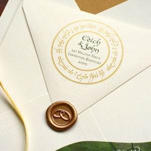 Op de afbeelding: Een witte envelop met een gouden waszegel en een ronde, gouden sticker met de tekst "Edith & John 547 Hilltop Drive Lexington, Kentucky 40503" in groene letters. De sticker is omgeven door een gouden cirkel met een inscriptie in het Elvish.