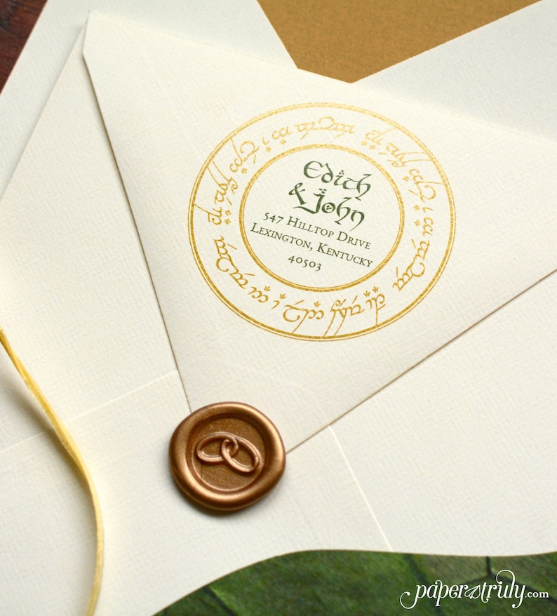 Op de afbeelding: Een witte envelop met een gouden waszegel en een ronde, gouden sticker met de tekst "Edith & John 547 Hilltop Drive Lexington, Kentucky 40503" in groene letters. De sticker is omgeven door een gouden cirkel met een inscriptie in het Elvish.