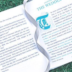 May include: A close-up of an open book with a silver ribbon bookmark. The book is titled "The Wedding" and features a blue and white illustration of a letter "T" with hearts. The text on the page describes a wedding ceremony and reception.