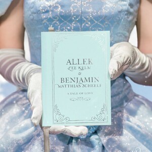 May include: A light blue book with silver lettering. The title is "AlleK Lee Kelm & Benjamin Matthias Scheele" and the subtitle is "A Tale of Love".