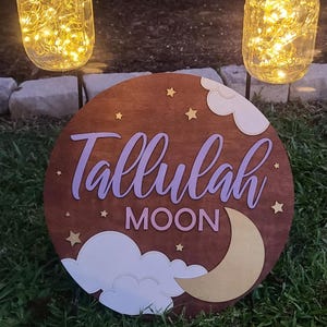 May include: A round wooden sign with the name "Tallulah Moon" in lavender script, adorned with stars, clouds, and a gold crescent moon. Two mason jars with fairy lights hang above the sign, creating a warm glow.